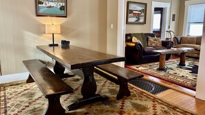 Un coin repas avec une table et des bancs en bois, un tapis à motifs et une vue sur le salon, où l’on aperçoit un canapé et un fauteuil.