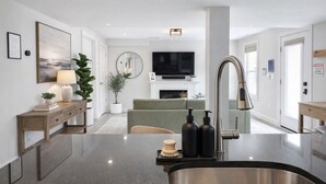 A modern kitchen with a stainless steel sink, a wooden console table, a green sofa, and a flat-screen TV.