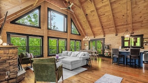 A spacious living area with a large window, a stone fireplace, a sofa, and a dining area with chairs and a table. - Arch Fun Cabin - Modern Cabin w/ Hot Tub, Games & Views (Stanton)