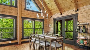 A wooden interior with a dining table and chairs, large windows, and a chandelier. - Arch Fun Cabin - Modern Cabin w/ Hot Tub, Games & Views (Stanton)