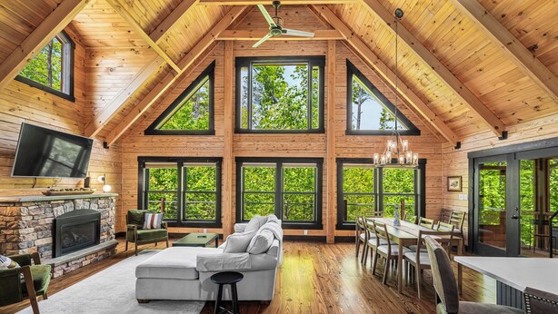 A spacious loft-style room with a stone fireplace, a large sectional sofa, a dining area with wooden tables and chairs, and large windows offering views of greenery. - Arch Fun Cabin - Modern Cabin w/ Hot Tub, Games & Views (Stanton)