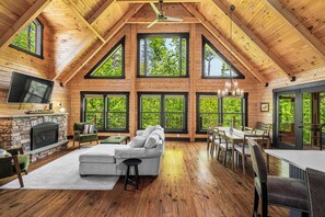 Een ruime loft-achtige kamer met een stenen openhaard, een grote modulaire bank, een eethoek met houten tafels en stoelen, en grote ramen met uitzicht op groen.