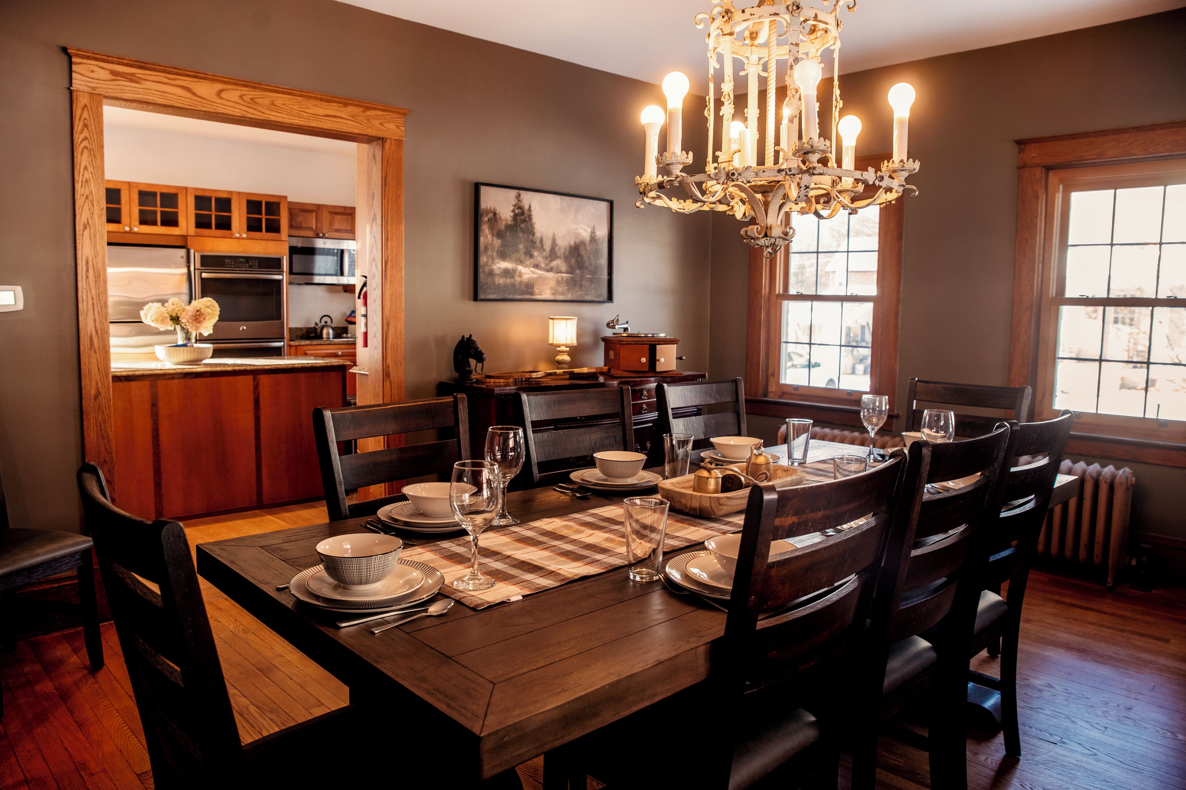 A dining area with a wooden table set for four, surrounded by chairs. A chandelier hangs above the table, and a painting is on the wall.