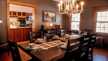 A dining area with a wooden table set for four, surrounded by chairs. A chandelier hangs above the table, and a painting is on the wall.