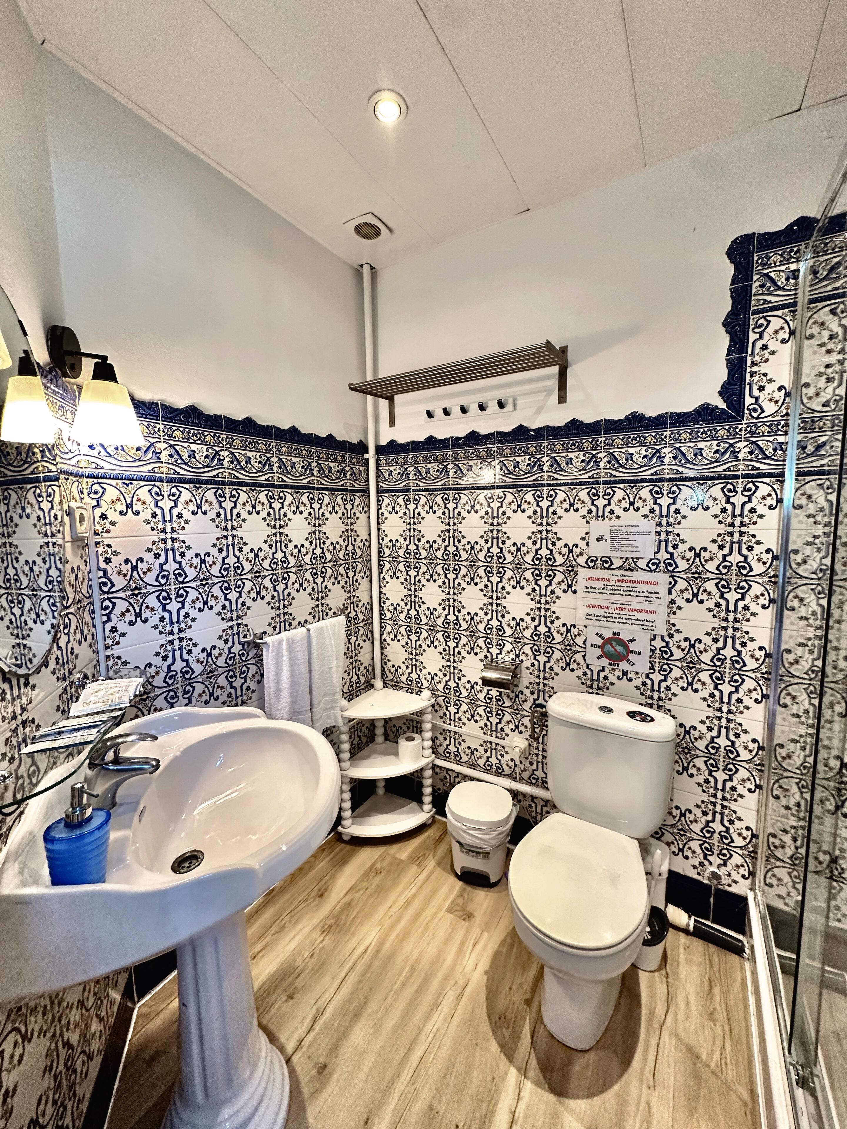 Une salle de bain avec un lavabo, des toilettes et une douche, le tout de couleur blanche. Les murs sont recouverts de carreaux aux motifs bleus et blancs. On y trouve un porte-serviettes et une étagère avec des articles de toilette.