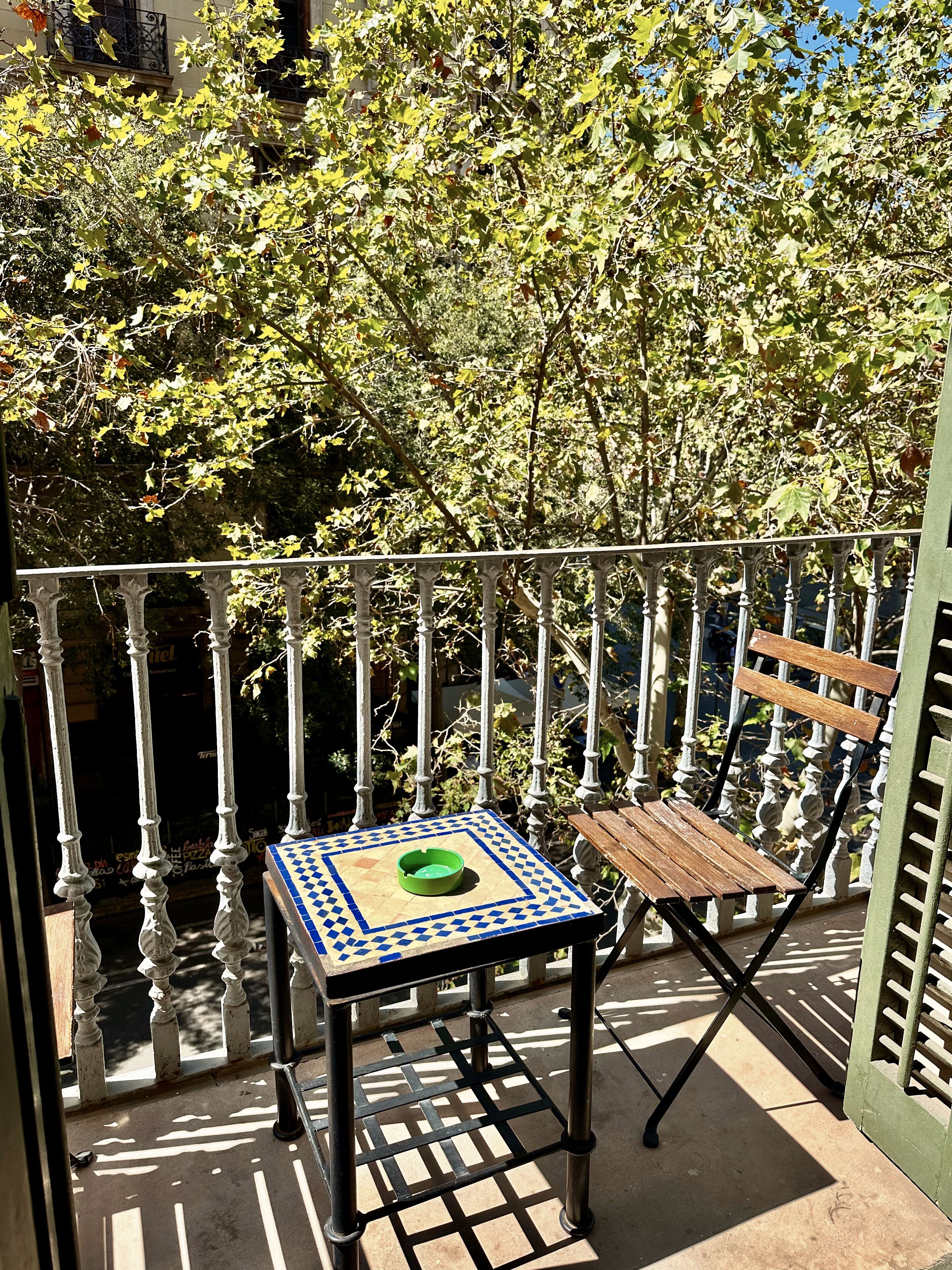 Un balcón con barandilla de metal, una mesa pequeña con un cuenco verde y un banco de madera.