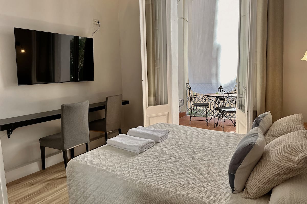 A hotel room with a bed, a desk, a chair, a TV, and a balcony with a table and chairs.