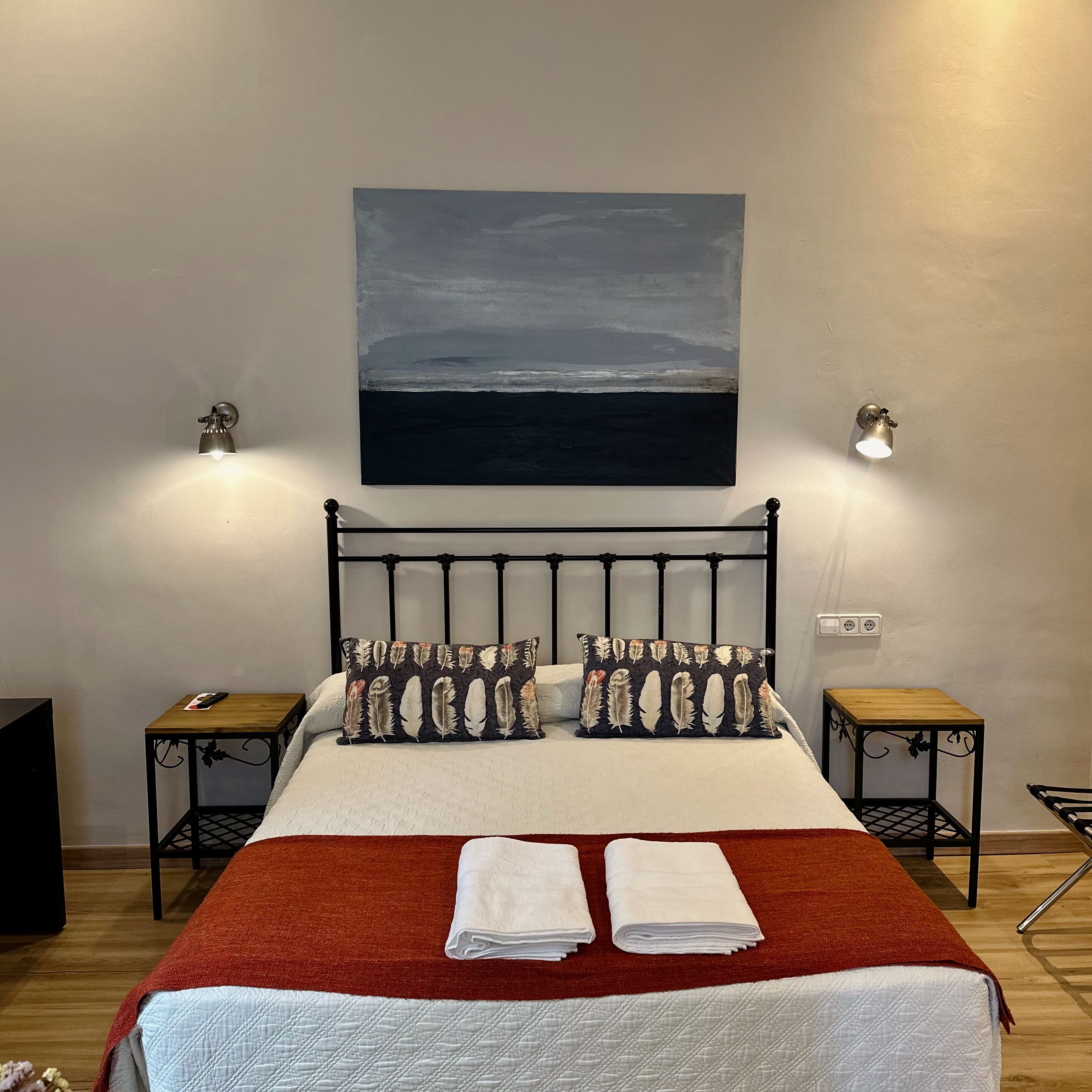 Una cama bien hecha con un edredón rojo, dos almohadas con estampado de plumas y un cuadro de un paisaje sobre ella. Hay dos mesitas de noche con lámparas, una mesita auxiliar de madera y un altavoz negro pequeño.