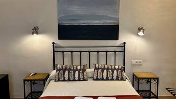 Una cama bien hecha con un edredón rojo, dos almohadas con estampado de plumas y un cuadro de un paisaje sobre ella. Hay dos mesitas de noche con lámparas, una mesita auxiliar de madera y un altavoz negro pequeño.