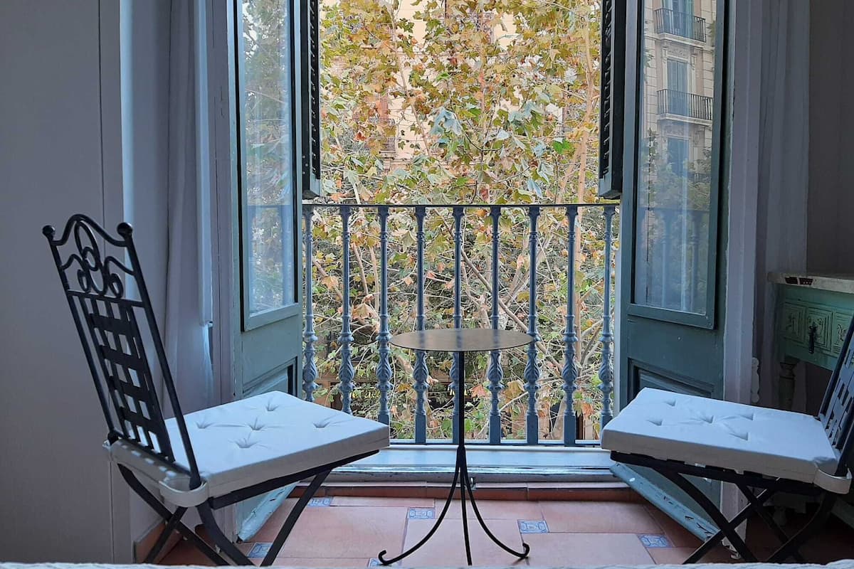 A balcony with two white cushioned chairs and a small table, overlooking a tree-lined street.