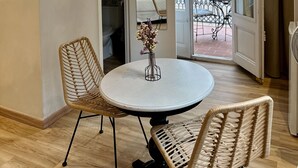 Un coin repas avec une table ronde, deux chaises en osier, un vase de fleurs et une vue sur un balcon doté d’une balustrade et d’une table à l’extérieur.