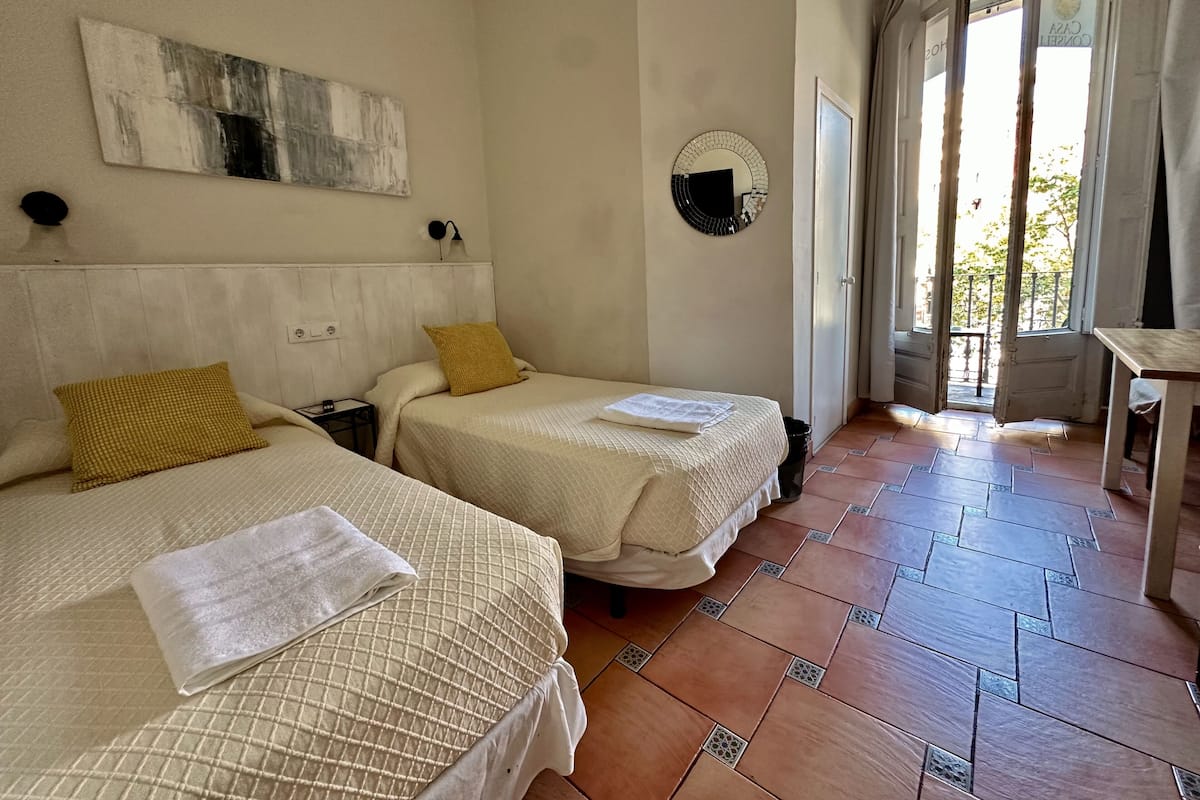 A hotel room with two beds, a large abstract painting, a round mirror, and a view of the outside through a door.