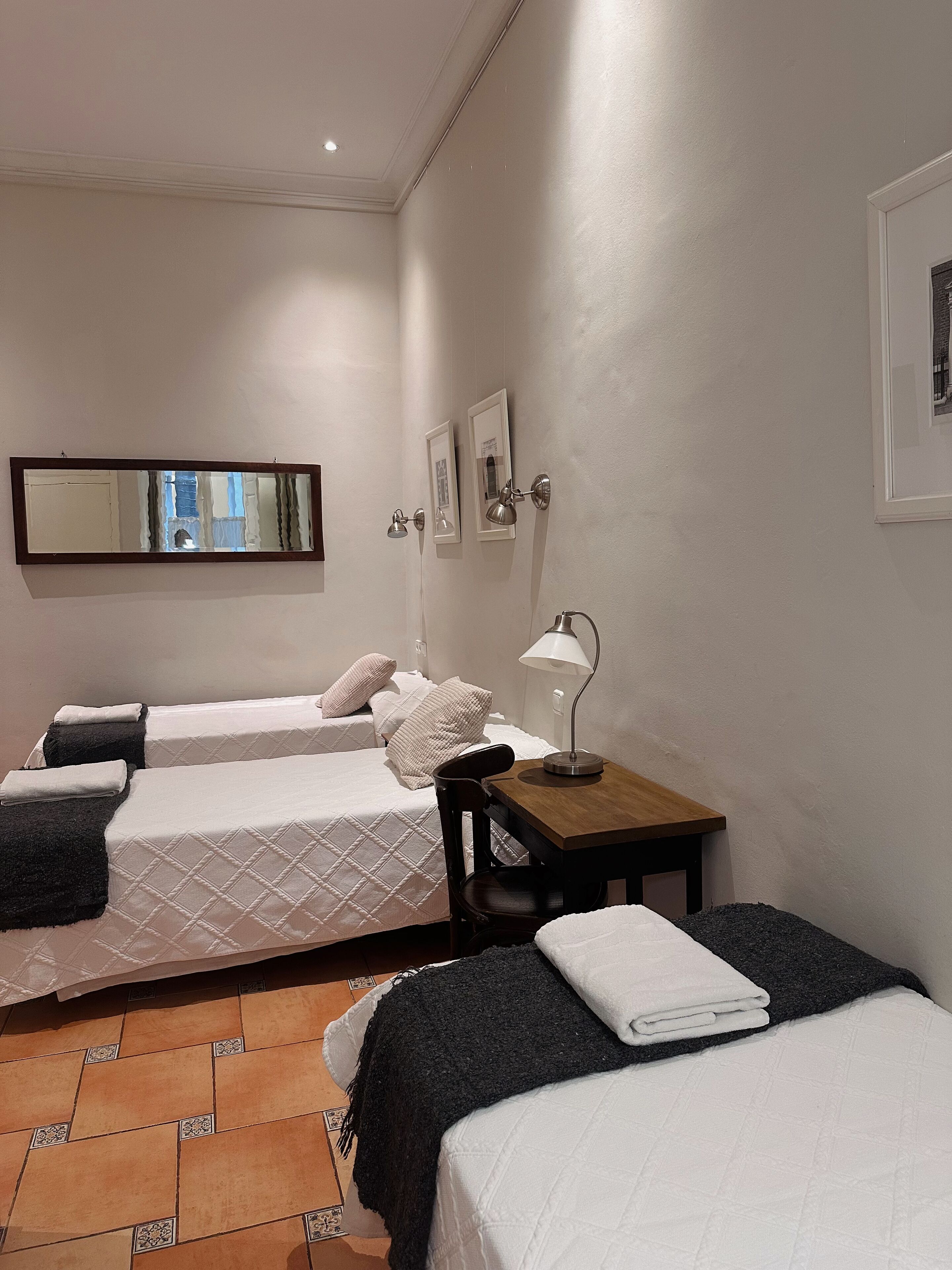 A hotel room with two beds, a small table with a lamp, a mirror, and framed pictures on the wall.