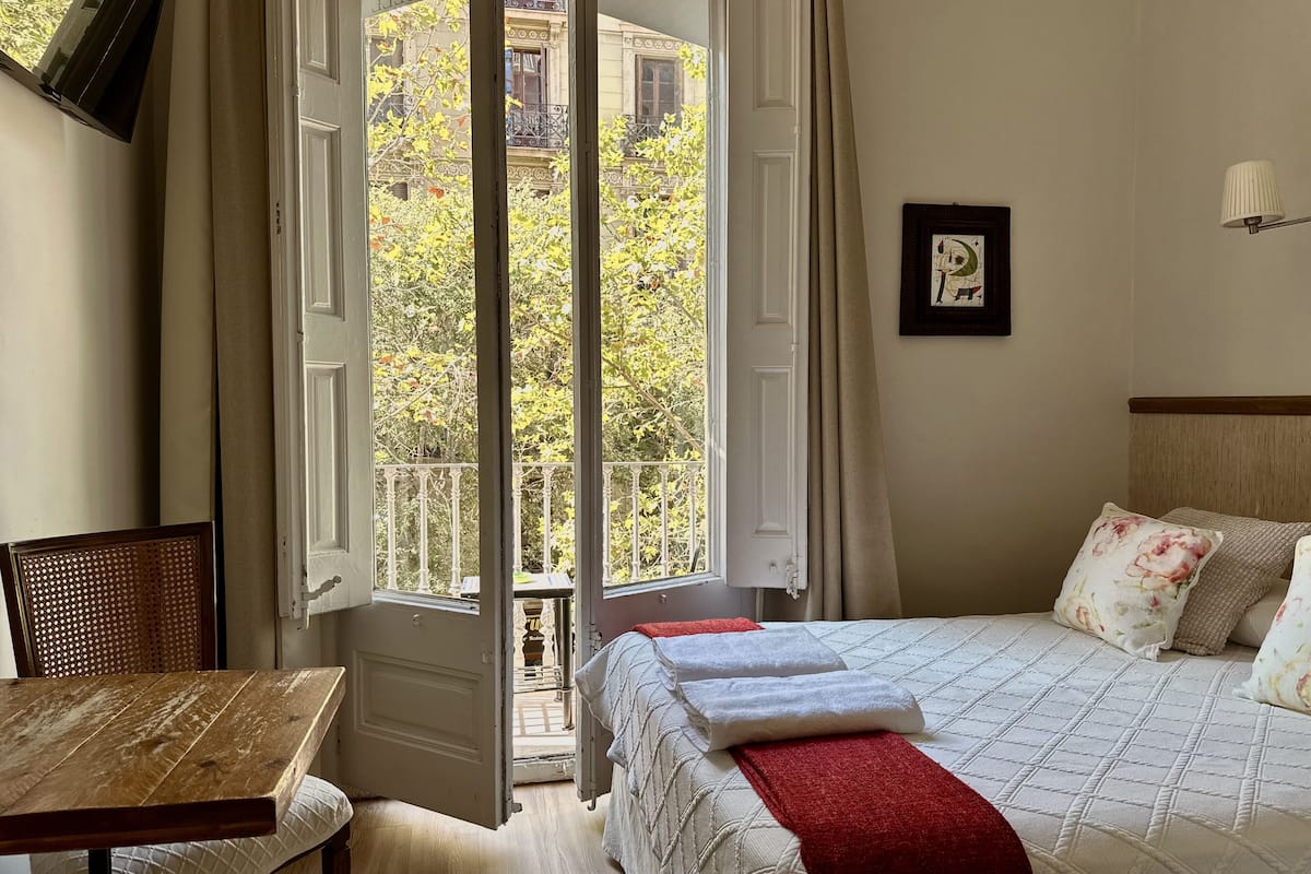 A hotel room with a bed, a table, a chair, and a balcony with a view of buildings and trees.