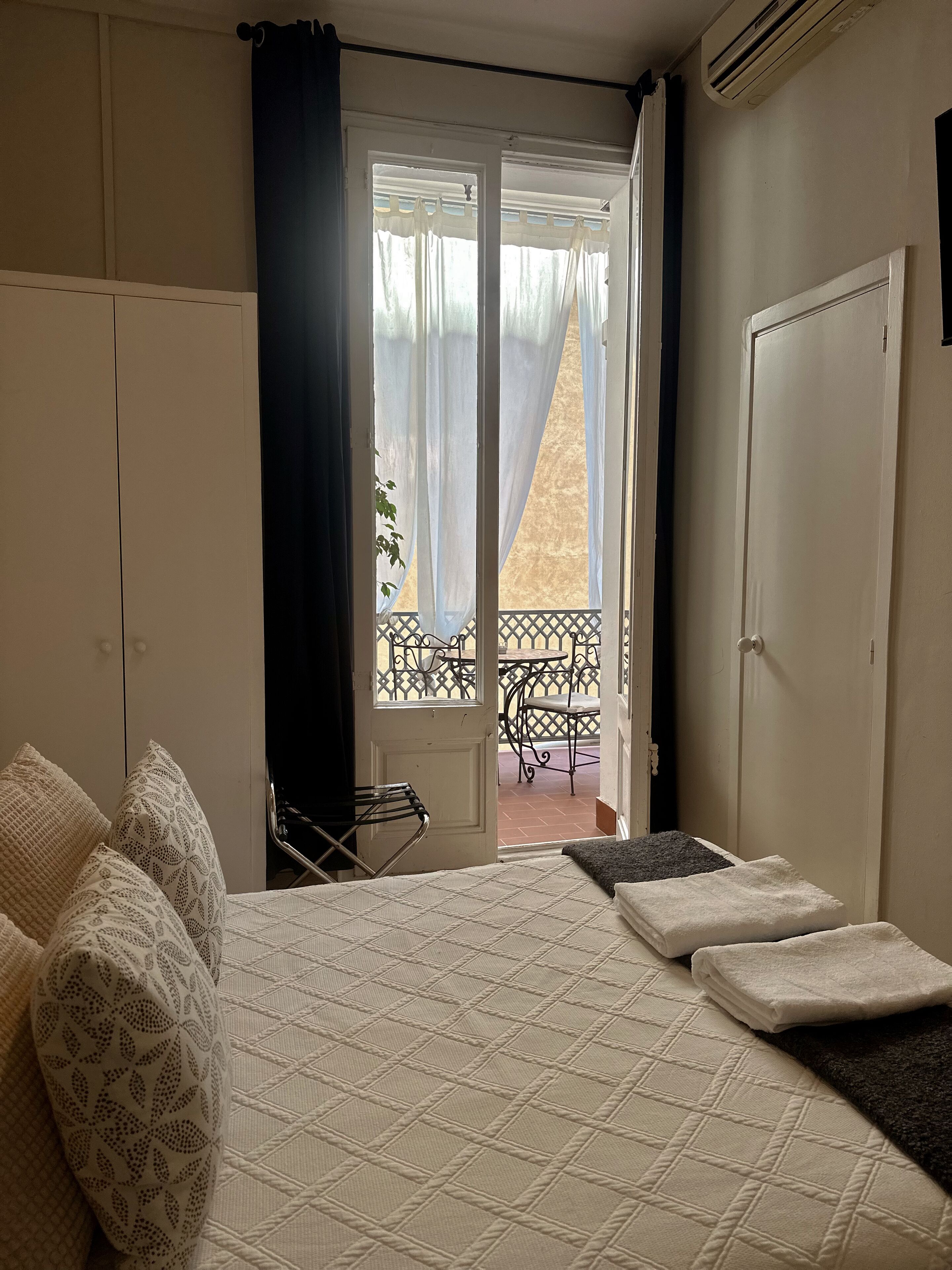 A bedroom with a bed, a window with curtains, a balcony with a table and chair, and a door.