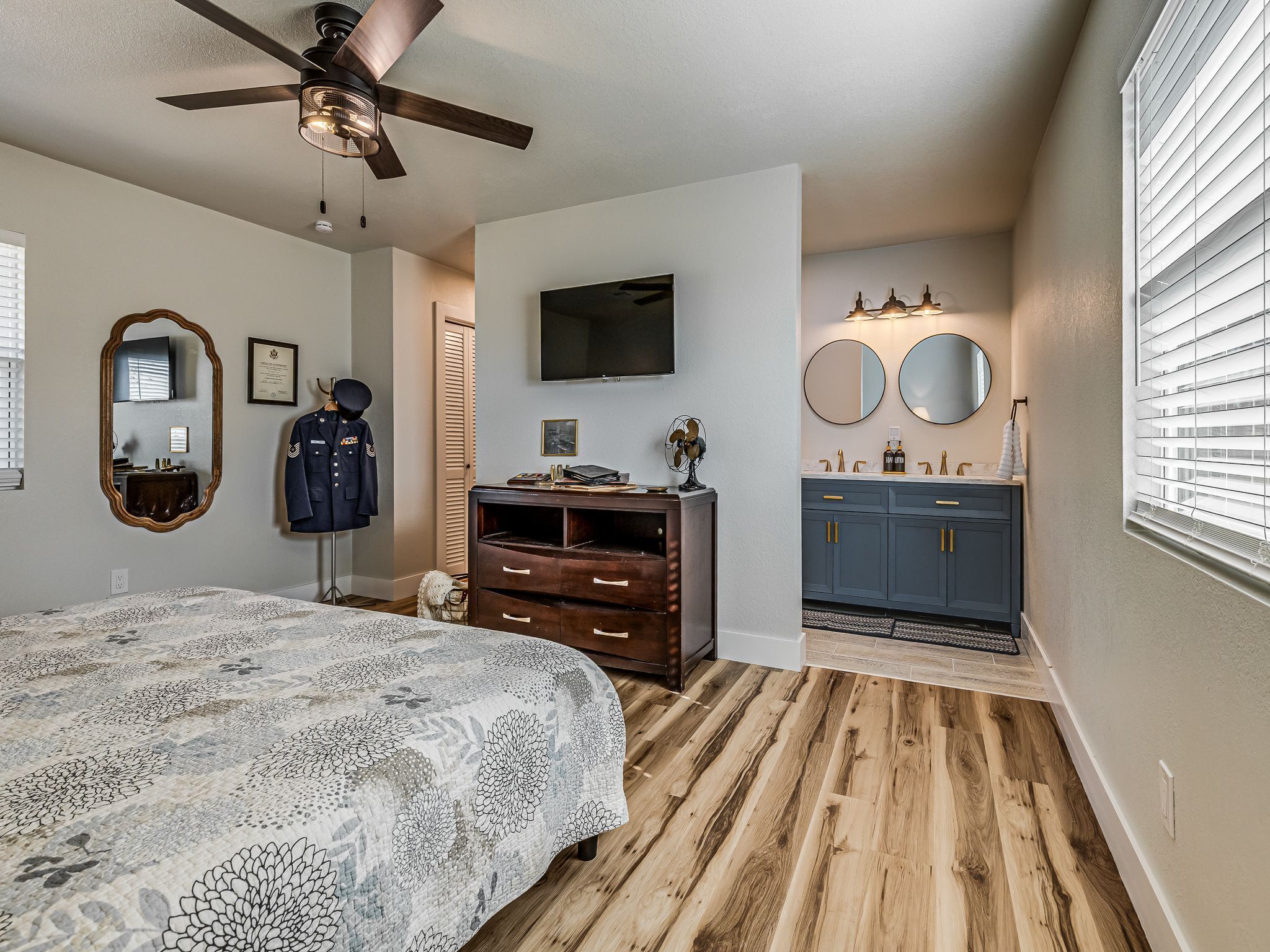 Une chambre à coucher comprenant un lit, un ventilateur de plafond, une commode, une coiffeuse avec des miroirs et un téléviseur fixé au mur.