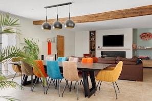 A modern living room with a dining area, wooden beams, a flat-screen TV, and colorful chairs.