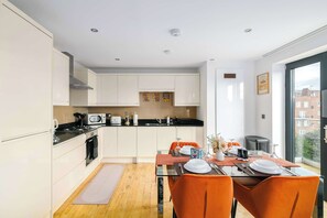 Une cuisine moderne avec des meubles blancs, un coin repas avec des chaises orange et une vue sur un quartier résidentiel par le biais d’une grande fenêtre.