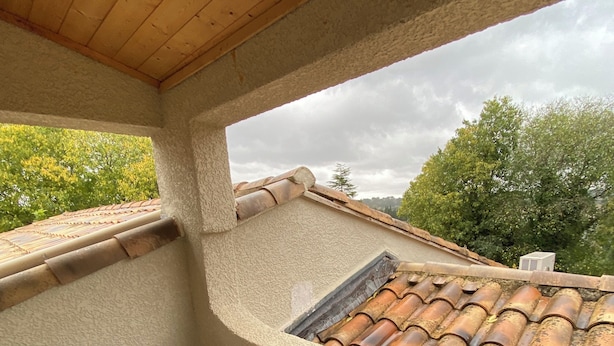 Een klein balkon met een betegeld dak, een houten tafel, twee rieten stoelen en uitzicht op bomen en een bewolkte lucht.