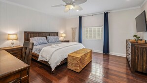 Een slaapkamer met een houten bed, nachtkastjes met lampen, een plafondventilator, een geweven bankje en een televisie aan de muur.