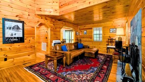 A log cabin interior with a wooden ceiling, a brown leather sofa with blue cushions, a red patterned rug, and a fireplace with a stone wall. - Private Ellijay Cabin Sleeps 6 - Loft, Stone Fireplace, Trails & Lake Nearby (Ellijay)