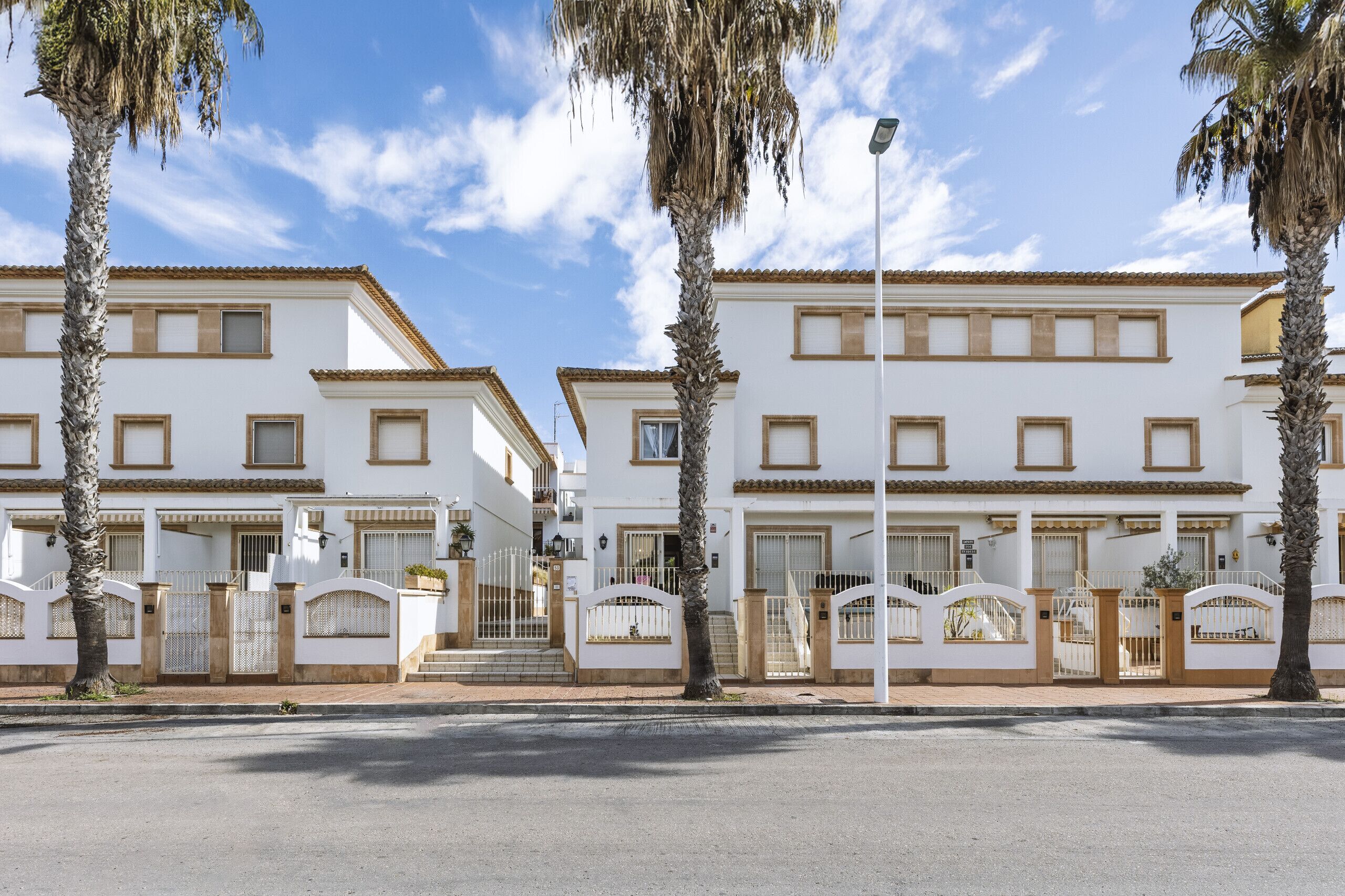 A row of white buildings with balconies, palm trees, and a clear sky.