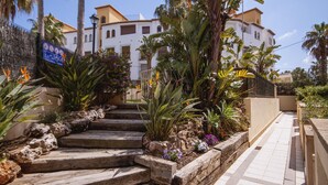 Un camino que conduce a un edificio de fachada blanca, rodeado de plantas tropicales y un muro de piedra.