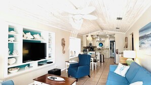 Un salon avec un téléviseur, des canapés bleus, une table basse et, au fond, un coin repas.