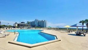 Une grande piscine extérieure, dotée de chaises longues et de parasols, entourée de bâtiments et surplombée par un ciel dégagé.