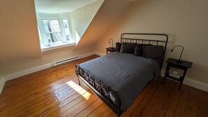 A bedroom with a sloped ceiling, a bed with a metal frame, a nightstand with a lamp, and a window with blinds. - Townhouse 3 at Historic Bauer Terrace (Halifax)