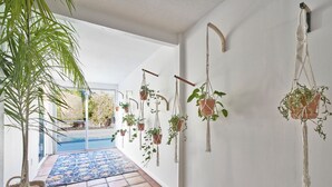 A hallway with hanging macramé plant hangers, a tiled floor, and a view of a pool. - Eclectic Bohemian Maximalist Pad with Pool and Spa (Desert Hot Springs)