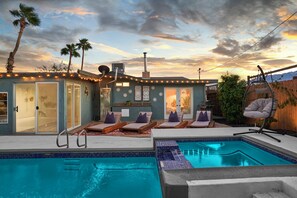 Un área de piscina con sillones de descanso y una piscina pequeña, rodeada por una casa con luces colgantes y palmeras.