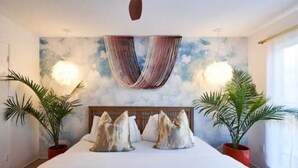 A bedroom with a bed, a headboard, two potted plants, and a wall with a cloud-like design. - Eclectic Bohemian Maximalist Pad with Pool and Spa (Desert Hot Springs)