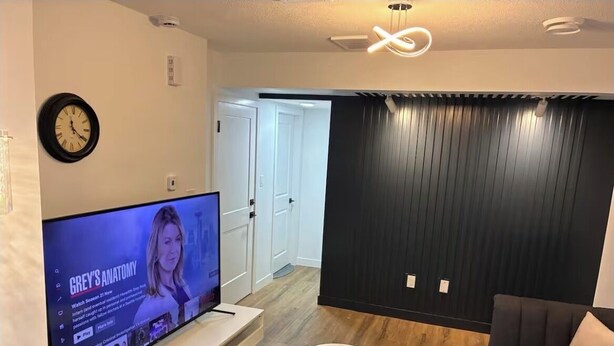 A modern living room with a flat-screen TV, a sofa, a coffee table, and a wall-mounted clock. - Home away from Home (Edmonton)