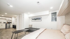 Et moderne soveværelse med en seng, et sengebord og et lille skrivebord med stol. Der er et tekøkken i baggrunden med skabe og en mikrobølgeovn.