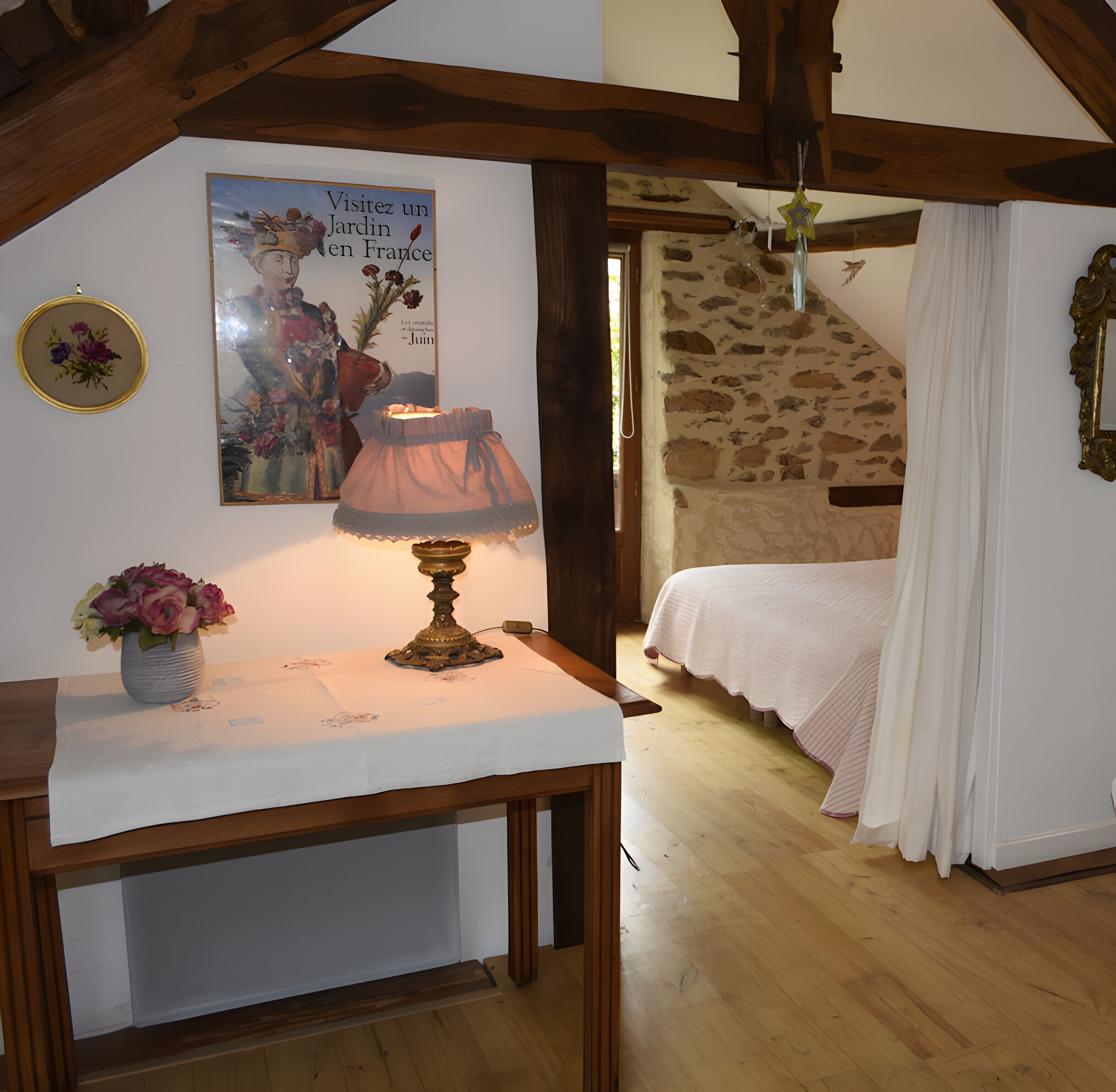 A bedroom with a wooden ceiling, a stone wall, a bed with white bedding, a bedside table with a lamp and flowers, and a poster on the wall.