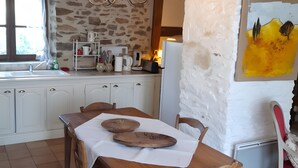 A rustic kitchen with a stone wall, wooden beams, a chandelier, a dining table with chairs, and a painting on the wall.