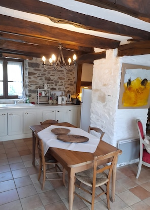 A rustic kitchen with a stone wall, wooden beams, a chandelier, a dining table with chairs, and a painting on the wall.