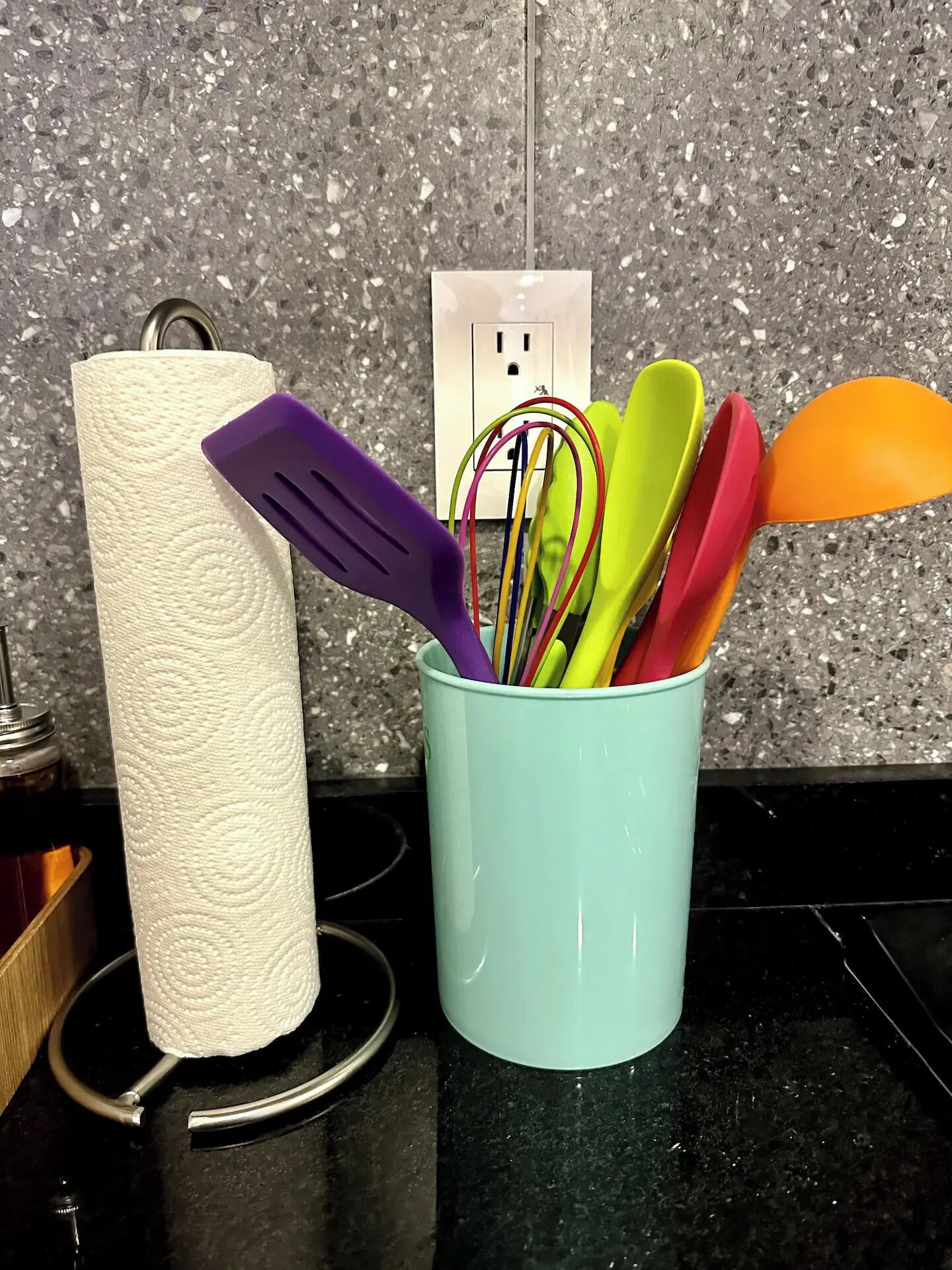 A kitchen counter with a paper towel holder, a cup of colorful utensils, and a granite backsplash.