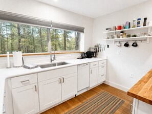 Une cuisine moderne avec des meubles blancs, un plan de travail en bois, un évier encastré et une fenêtre donnant sur la forêt.