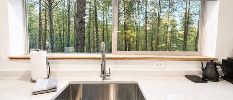 Une cuisine équipée d’un évier en acier inoxydable, d’une fenêtre donnant sur les arbres, d’une bouilloire et d’un égouttoir à vaisselle.