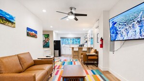 Un salon moderne avec un canapé, une table basse et un tapis rayé aux couleurs vives. Un téléviseur est fixé au mur et un ventilateur de plafond est installé.