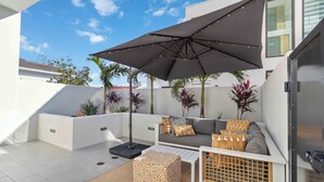 Un patio moderno al aire libre con un sofá, cojines, un taburete estampado y una sombrilla grande.