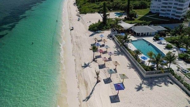 Vista aérea de un complejo turístico en la playa, con un mar turquesa transparente, una playa de arena bordeada de sombrillas y una zona de piscina junto a un edificio.