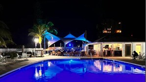 Área de piscina nocturna con bar, asientos tipo lounge, palmeras y una piscina cristalina.