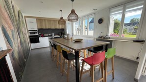 Moderni keittiö, jossa on puinen keittiösaareke, baarituolit, riippuvalaisimet ja suuret ikkunat, joista avautuu näkymä ulos.