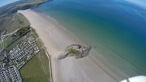 Vue aérienne d’une zone côtière comprenant une plage de sable, un petit promontoire rocheux et une zone résidentielle avec des maisons et des routes.