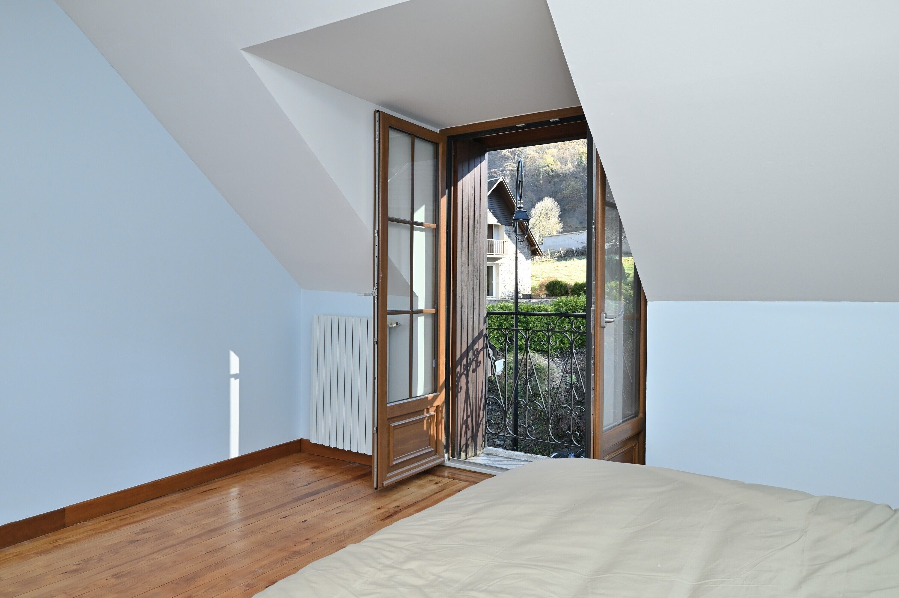 A bedroom with a sloped ceiling, a bed, a radiator, and a balcony with a view of a house and greenery.