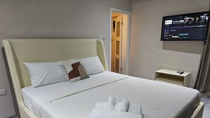 Una cama bien hecha con ropa blanca y un cojín marrón decorativo, una toalla blanca doblada sobre la cama, una mesita de noche con un teléfono y un televisor montado en la pared mostrando YouTube.