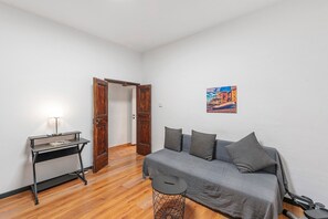 Une pièce avec une porte en bois, un canapé gris avec des coussins, une petite table ronde et une lampe posée sur un pied métallique.